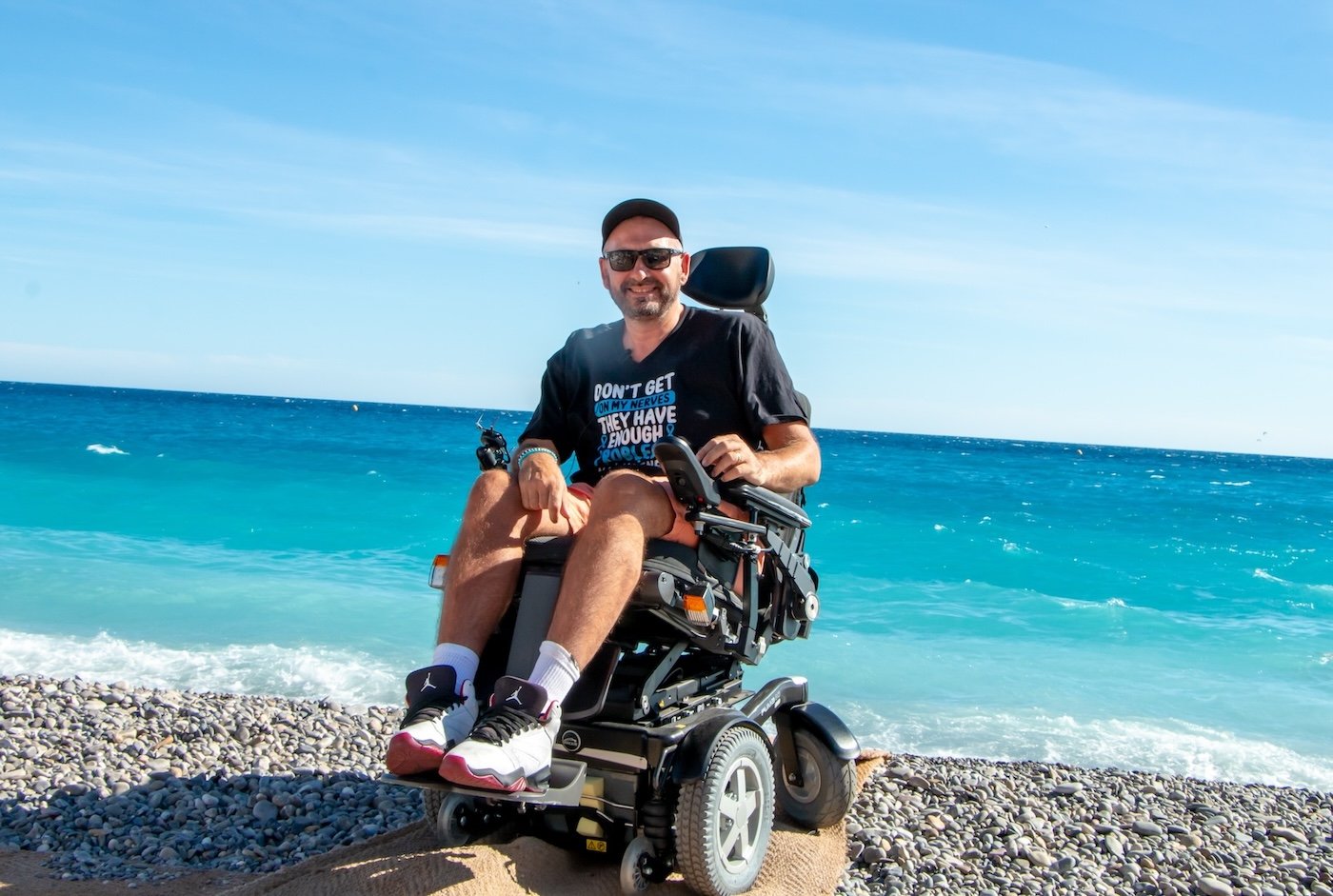 En smilende mann i elektrisk rullestol sitter på stranden. Han har på seg caps, solbriller og en svart t-skjorte med. I bakgrunnen ser man et turkist hav med hvite bølger som slår mot den steinete stranden. Himmelen er klar og blå. Bildet utstråler en avslappet ferie-atmosfære 